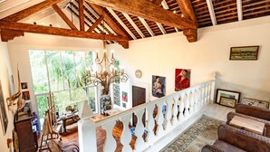 Une pièce spacieuse avec un plafond haut, des poutres en bois, un lustre, un balcon et une vue sur la verdure environnante.