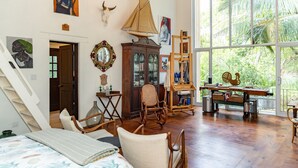 Une pièce comprenant un lit, des chaises en bois, un bureau avec un ordinateur, une grande fenêtre donnant sur un paysage verdoyant et divers éléments de décoration murale.