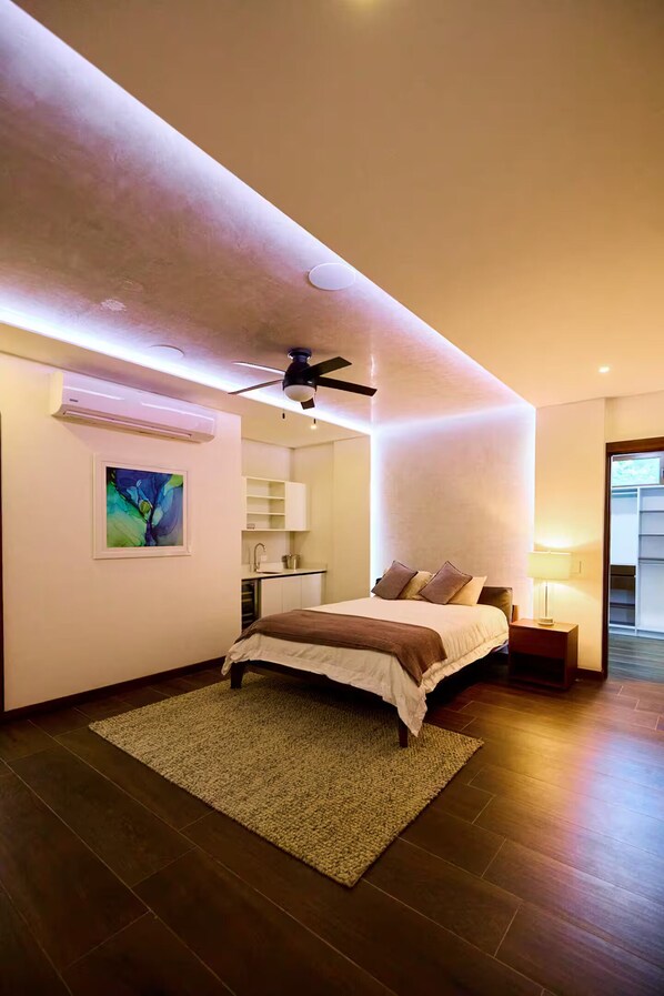 Quarto moderno com cama, ventilador de teto, quadro e cozinha compacta.