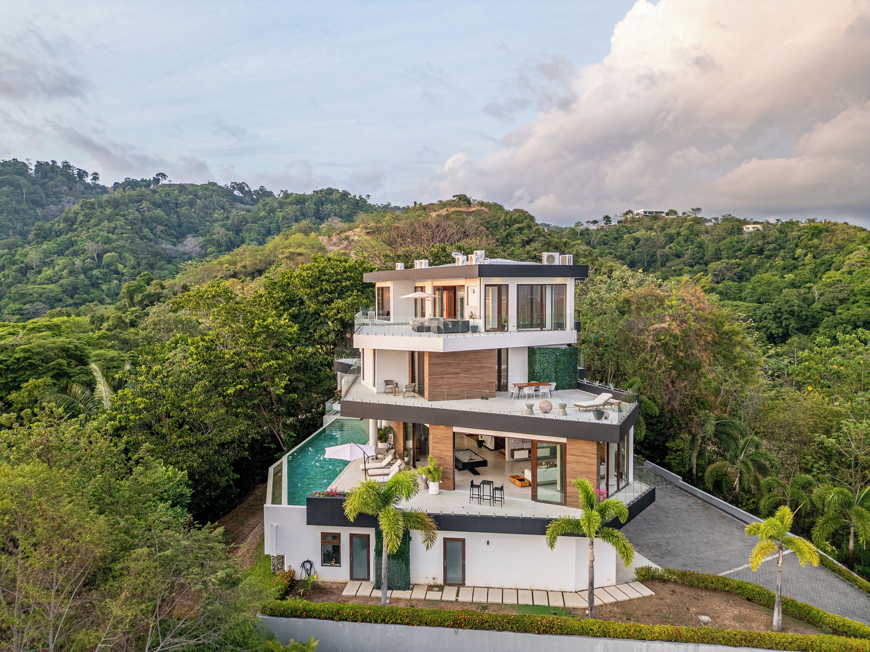 A modern multi-story house with a rooftop pool, surrounded by lush greenery and hills.