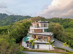 Uma casa moderna de vários andares com piscina no terraço, rodeada por vegetação exuberante e colinas.