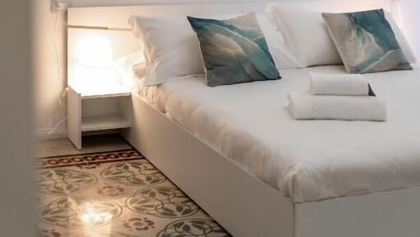A neatly made bed with pillows and a nightstand, a patterned rug, and a wall-mounted picture of a cityscape. - La Casa di Lidia - Vatican City  (Rome)