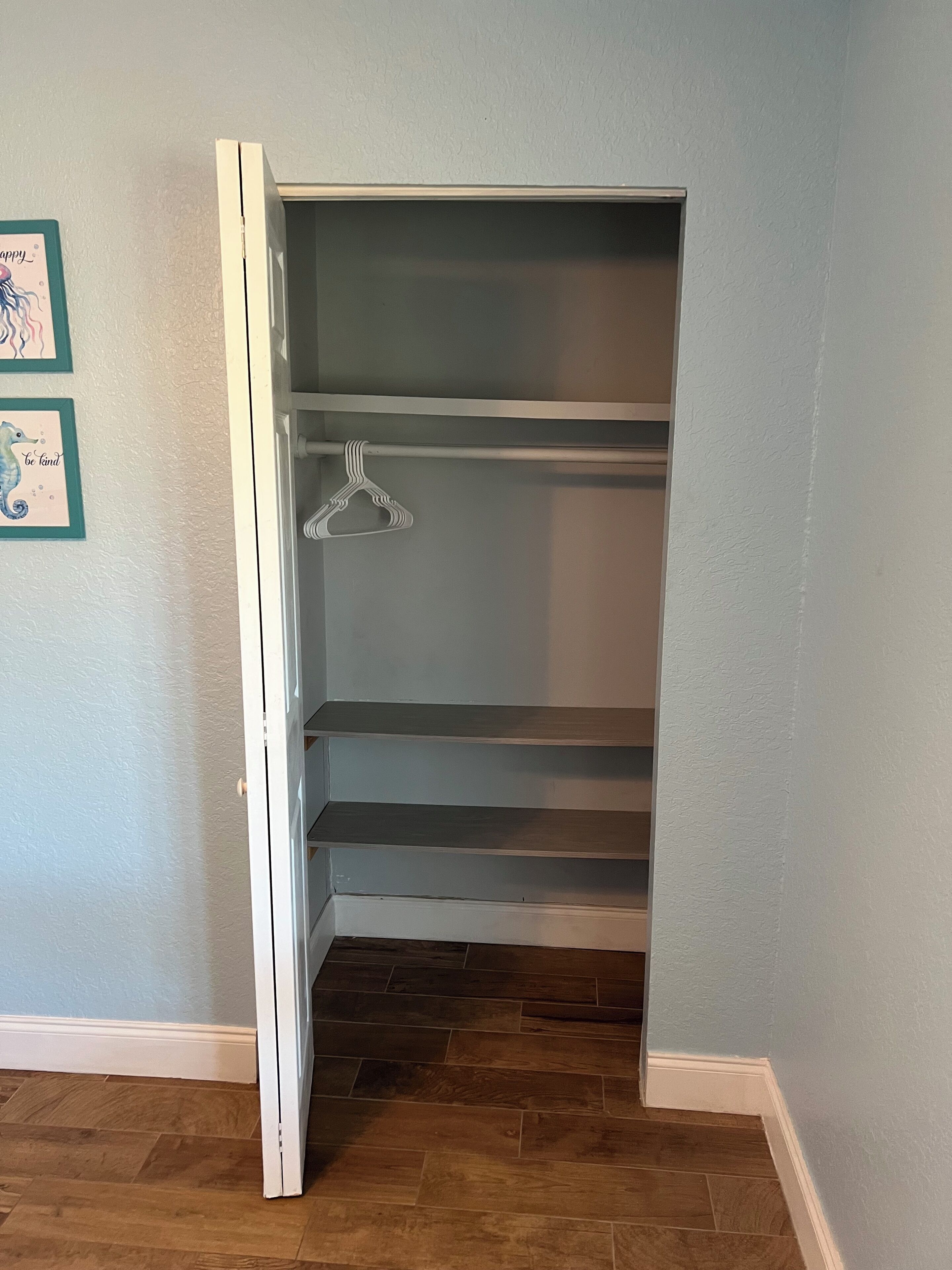 A closet with open doors, wooden flooring, and a wall with two framed pictures.