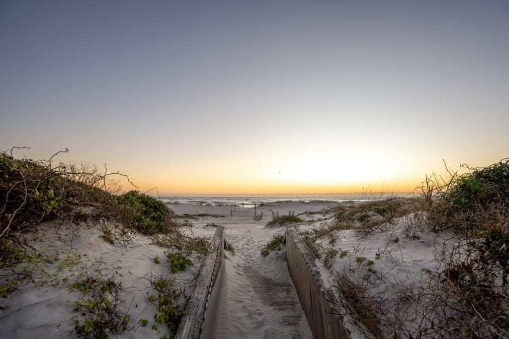 A sandy beach path leading to the ocean with a wooden fence on either side.