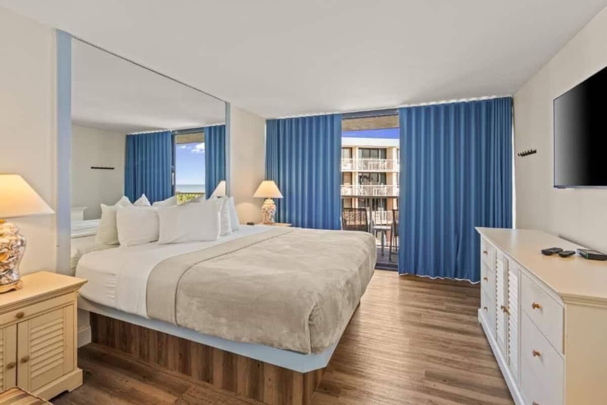 Habitación de hotel con cama, mesitas de noche, televisor, cómoda y balcón con cortinas.