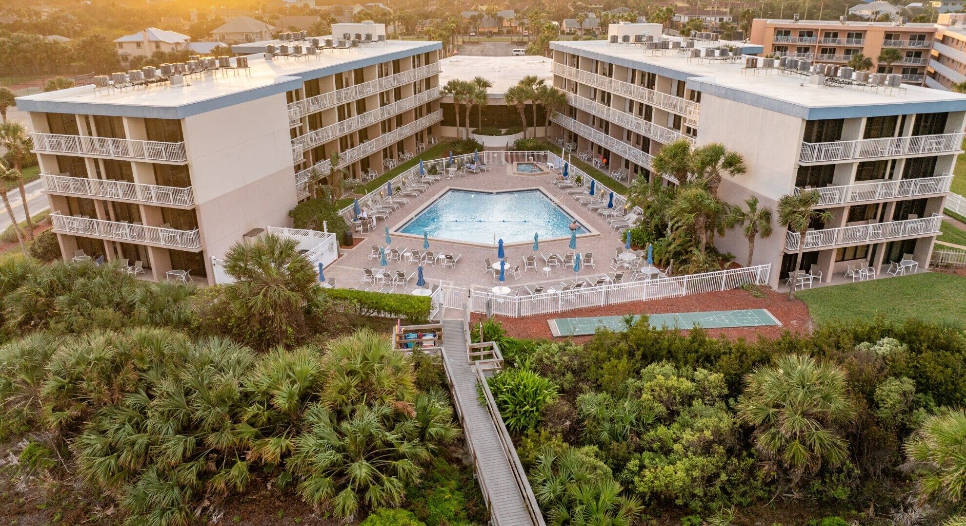 A hotel with a central pool surrounded by lounge chairs and umbrellas, multiple balconies, and lush greenery.