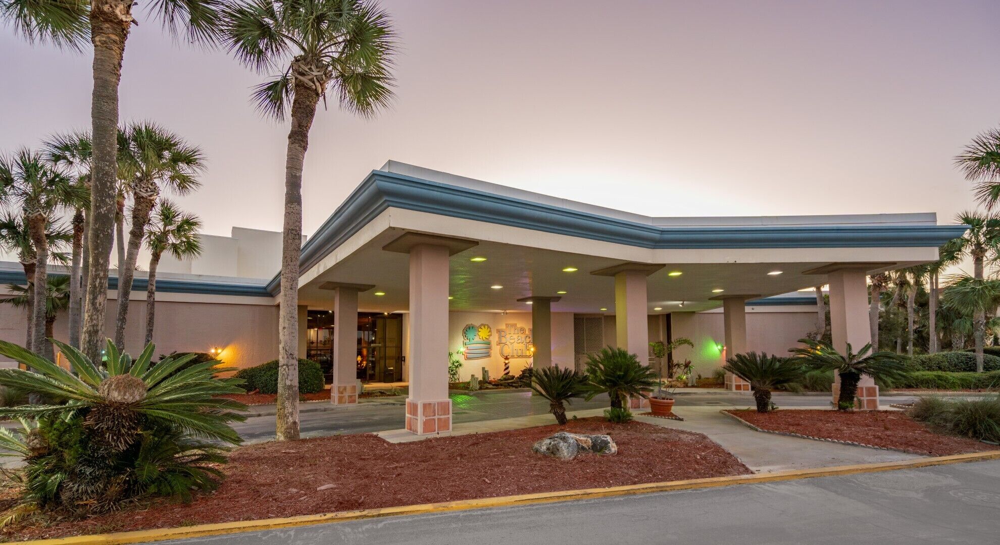 Een hotelentree met een overdekt terras, palmbomen en een bord met 'The Beach Club'.