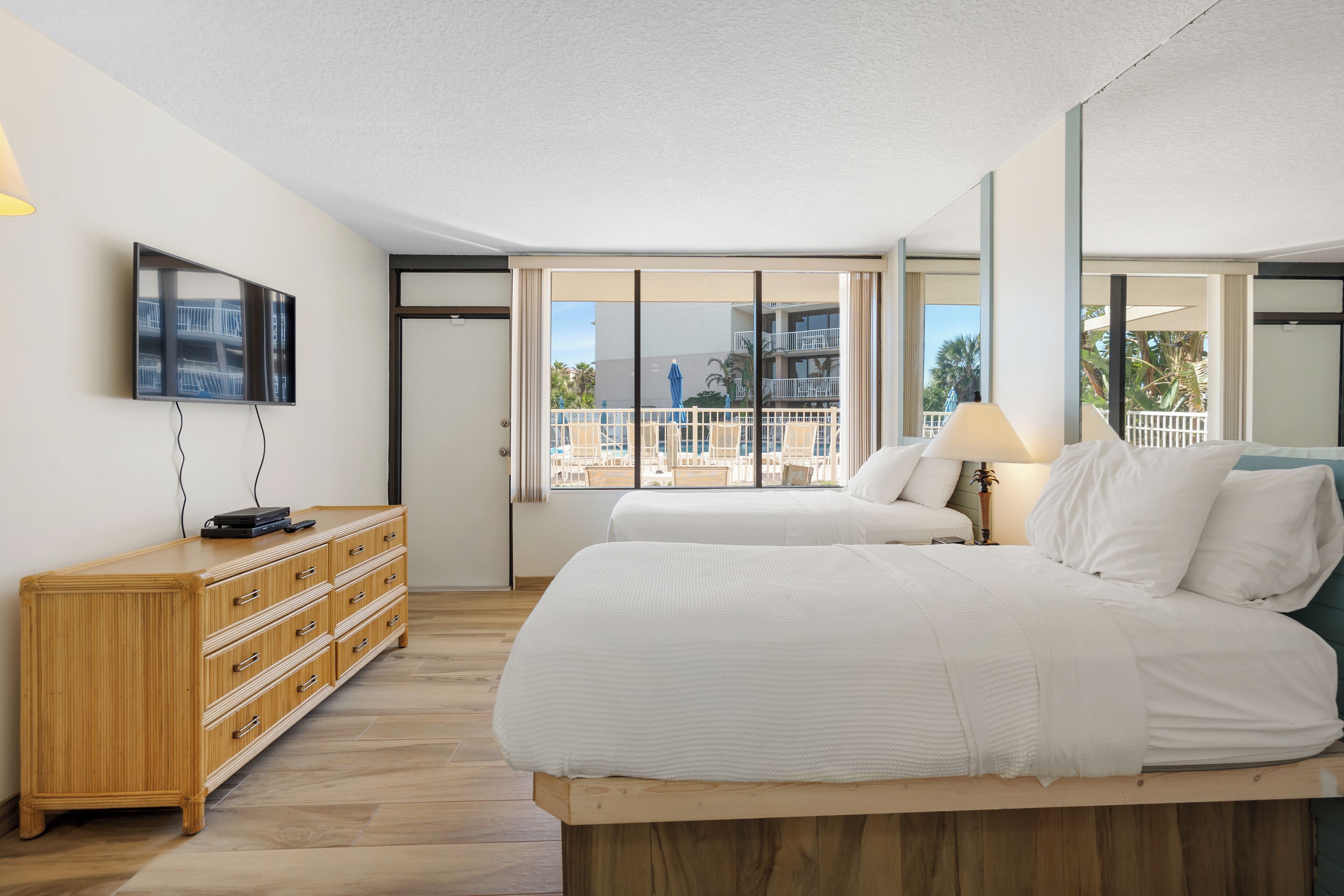 Ein Hotelzimmer mit einem Bett, einer Holzkommode, einem an der Wand montierten Fernseher und großen Fenstern mit Blick auf die Stadt.