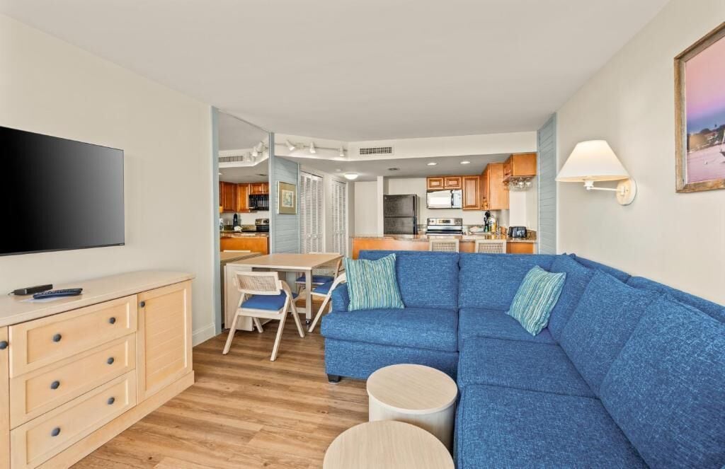 A modern living room with a blue sofa, a TV, a dining area, and a kitchen in the background.