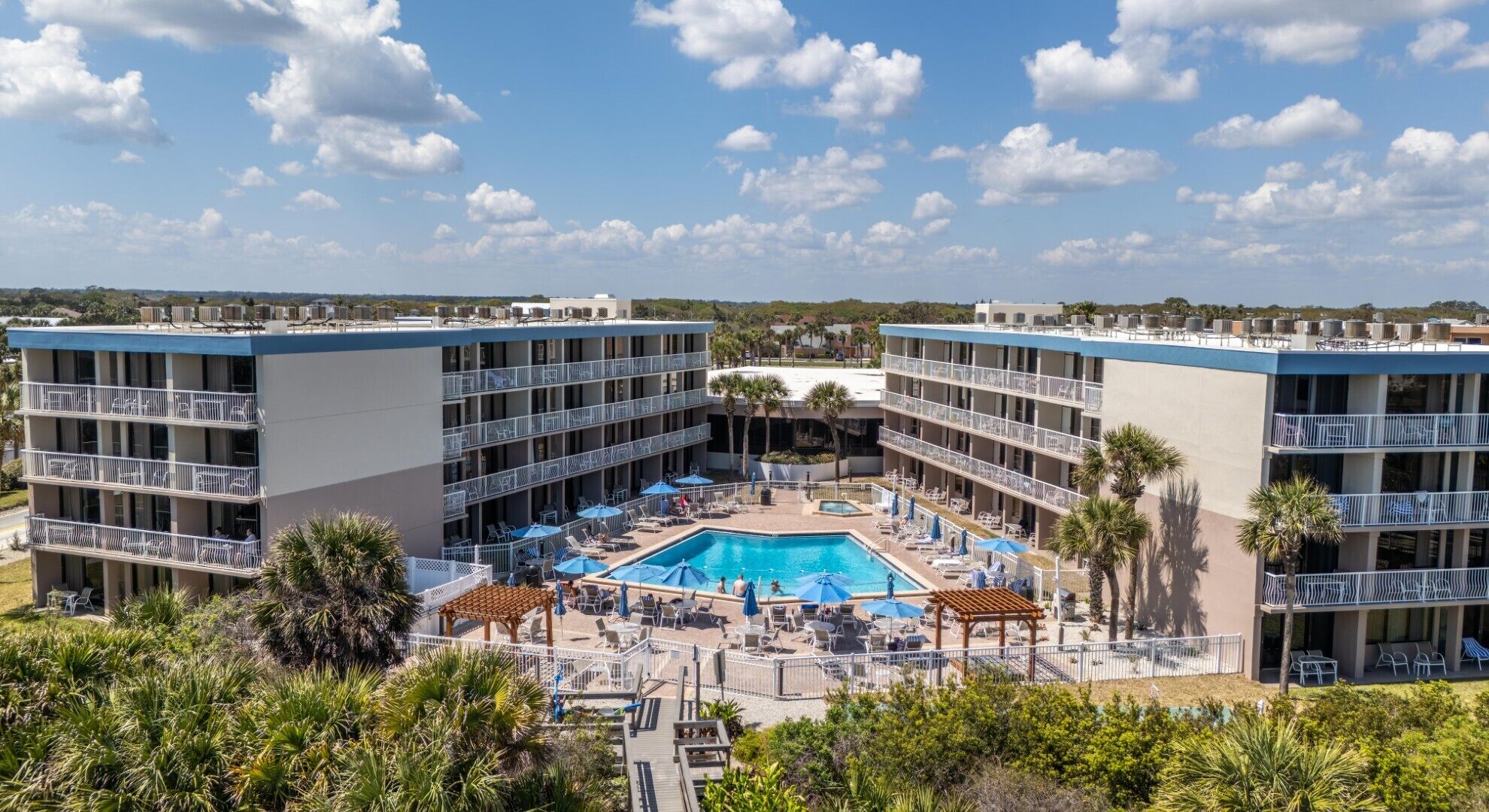 Ein Hotel mit einem zentralen Pool, umgeben von Liegestühlen und Sonnenschirmen, mehreren Balkonen und freiem Blick auf die umliegende Landschaft.