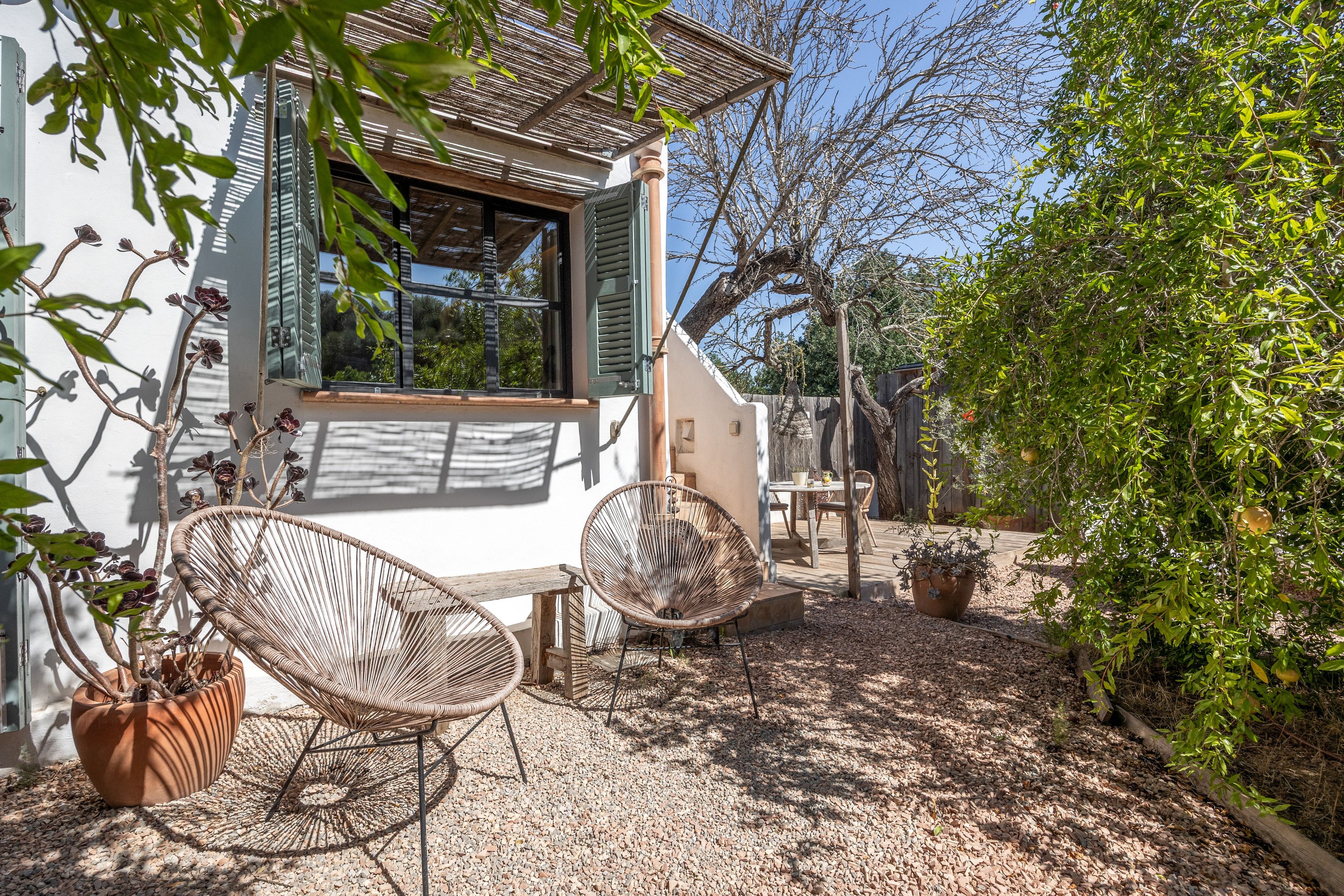 Een huis met een patio, voorzien van rieten stoelen en een tafel, omringd door een tuin met bomen en een houten omheining.