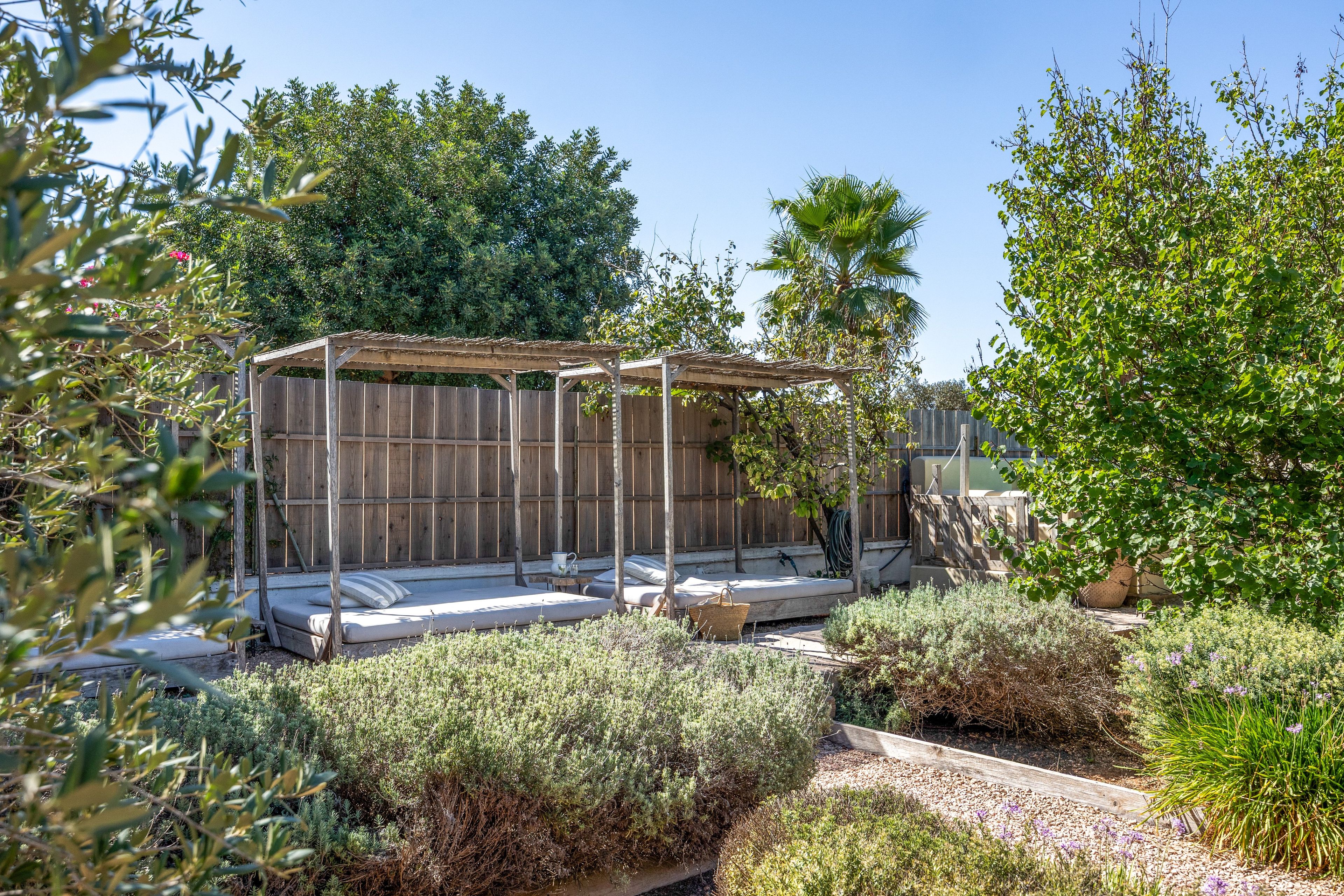 Een tuin met een houten pergola, een zithoek met kussens, en diverse planten.