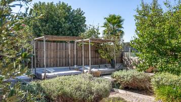 Een tuin met een houten pergola, een zithoek met kussens, en diverse planten.