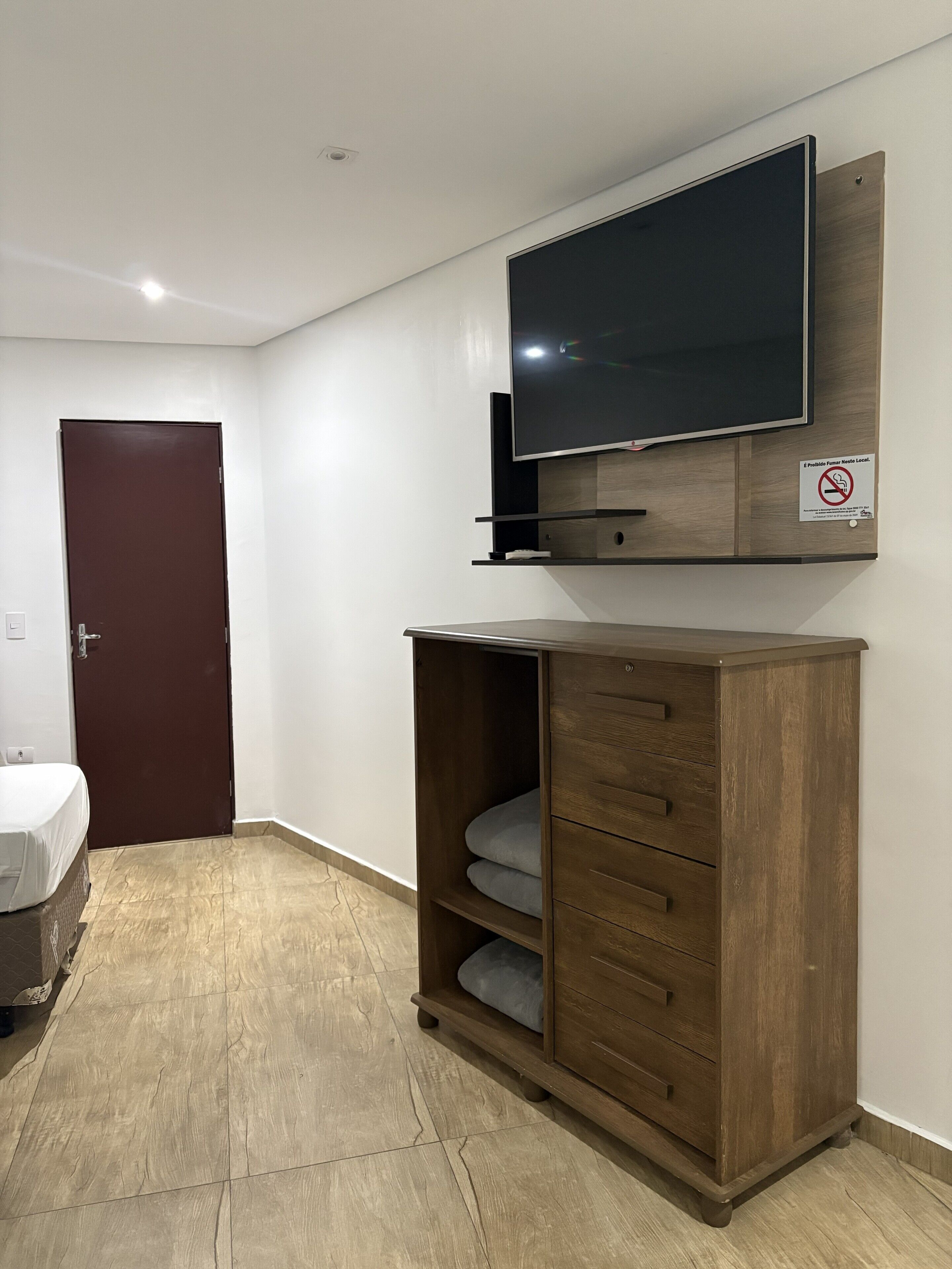 Une chambre d’hôtel avec un téléviseur à écran plat fixé à un meuble en bois, une porte fermée et un lit recouvert d’une couverture blanche.