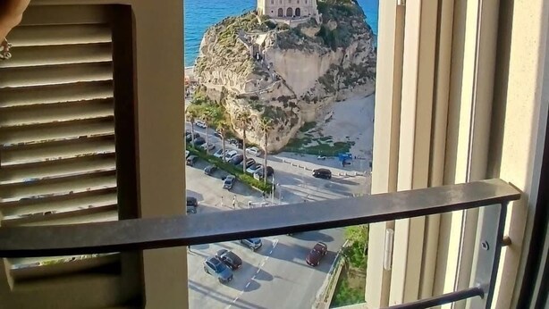 A balcony with a view of a coastal building on a cliff, a parking lot, and a clear sky. - TROPEA VETERA Luxury Home - Oven House 1638 (Tropea)