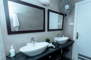 Un baño con dos lavamanos, una toalla colgada en el espejo de la izquierda y un ventilador a la derecha.