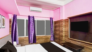 A hotel room with a bed, a TV, a window with purple curtains, and an air conditioning unit. - Hotel O Pradhan Nagar Prince Guest House (Siliguri)