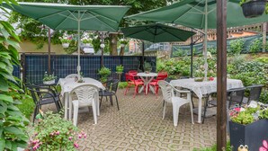 Ein Terrassenbereich im Freien mit Tischen und Stühlen, beschattet von grünen Sonnenschirmen, umgeben von Blumentöpfen und Blumen.