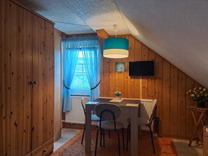 A room with wooden walls, a dining table with chairs, a ceiling fan, a TV mounted on the wall, and a window with curtains.