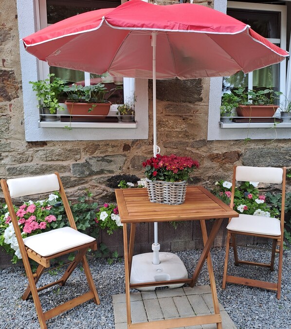 A wooden table with a white cushion, two wooden chairs, a red umbrella, and potted plants on a stone wall.