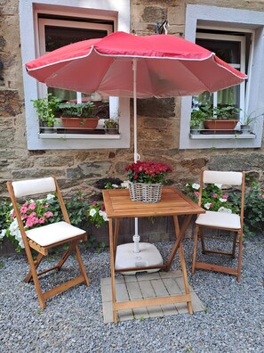 A wooden table and two chairs with a red umbrella, surrounded by potted plants and flowers.