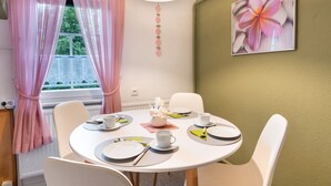 Et spiseområde med et rundt bord dækket til fire personer, hvide stole og et vindue med lyserøde gardiner.