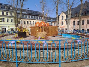 Une place publique avec une fontaine, une structure en bois agrémentée de décorations colorées et des bâtiments en arrière-plan.