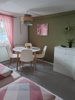 Une pièce avec une table de salle à manger ronde, des chaises blanches, une commode blanche, une plante en pot et une fenêtre ornée de rideaux légers.