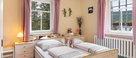 Ein Schlafzimmer mit Doppelbett, Nachttischen, einem Schminktisch, einem Fenster mit Vorhängen, einer Heizung und einer kleinen Pflanze an der Wand.