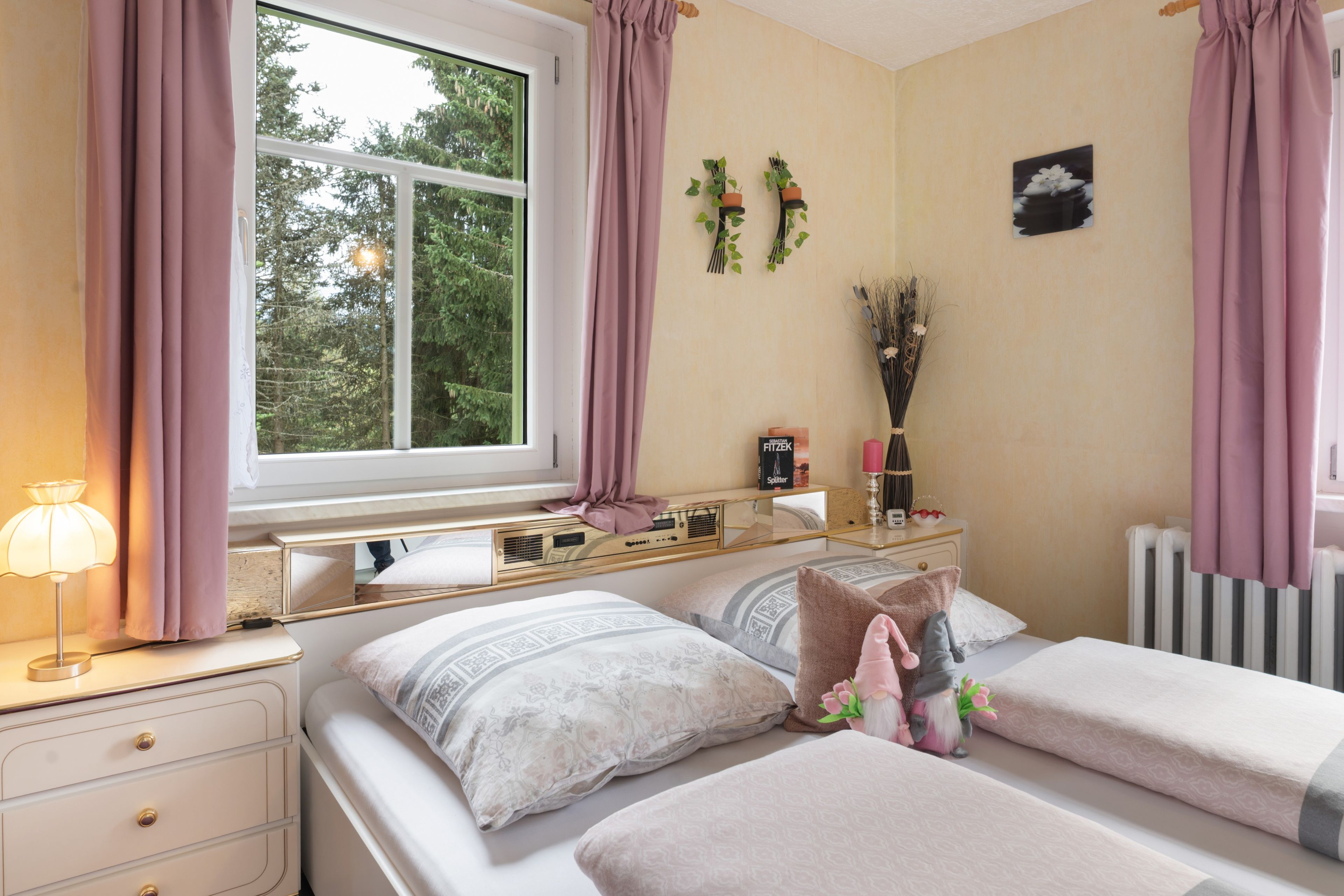 A bedroom with two beds, a bedside table with a lamp, a dresser, a window with curtains, and a wall-mounted plant.