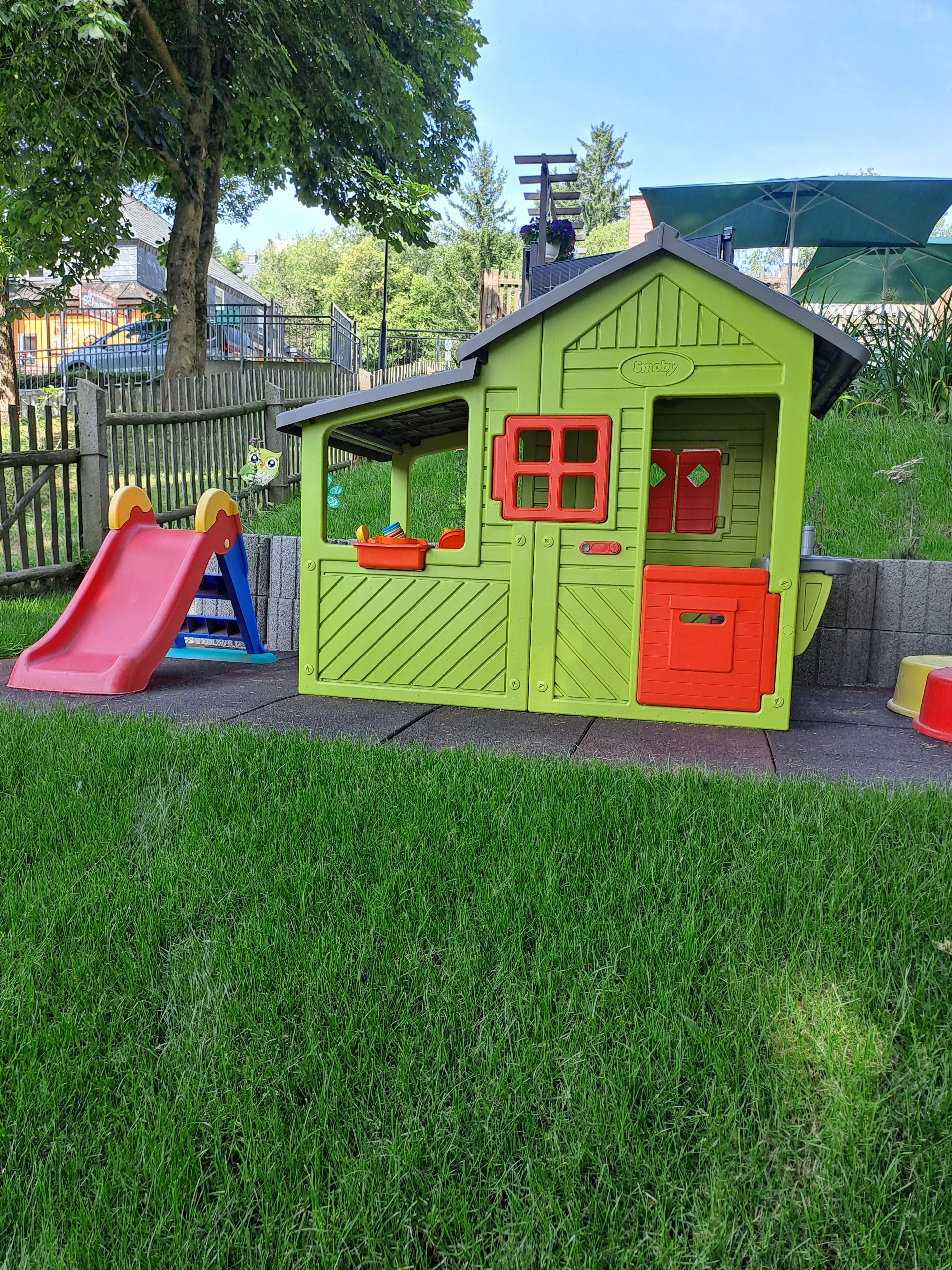 A children's playhouse with a slide, located in a grassy area with a fence and trees in the background.
