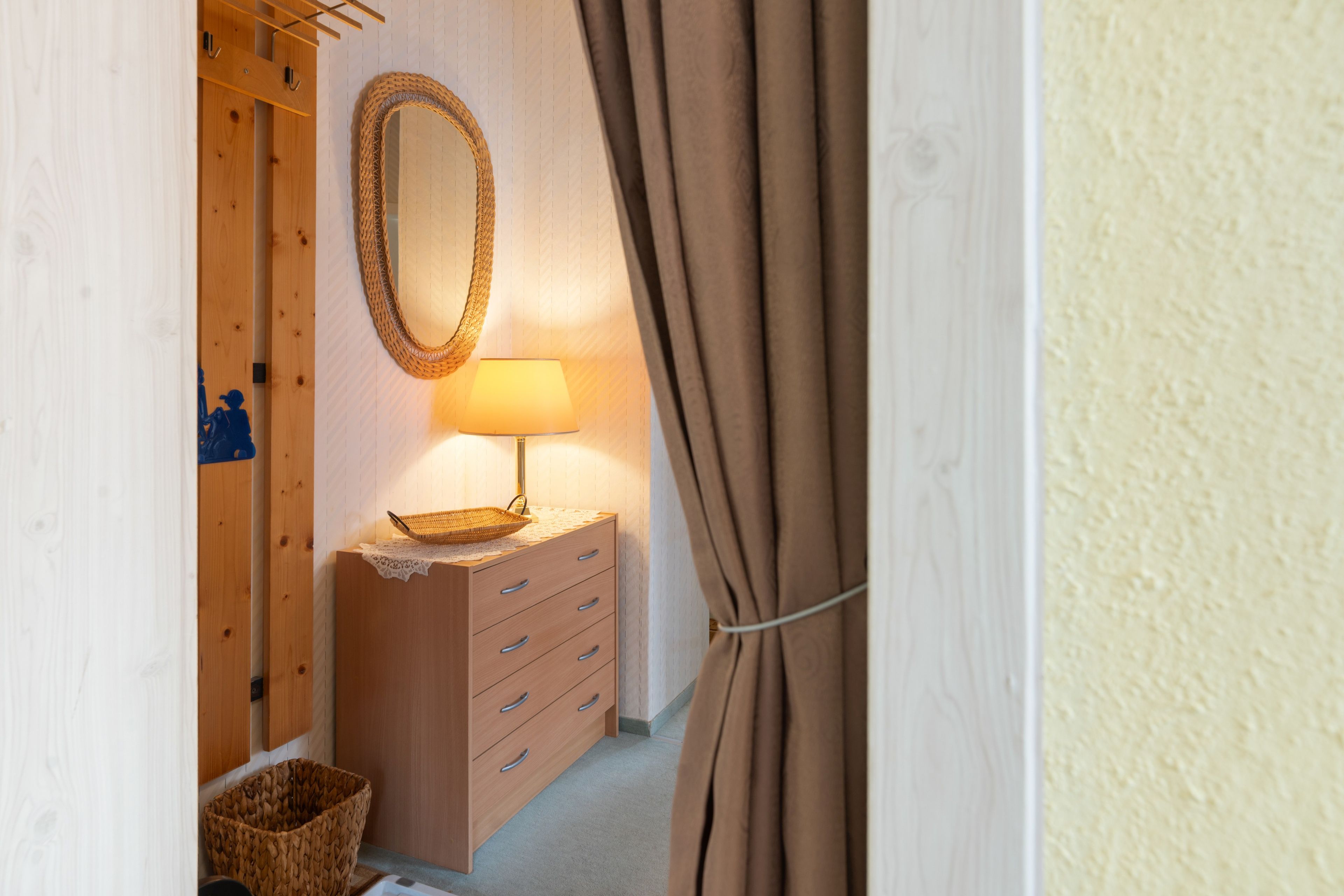 A wooden dresser with drawers and a lamp, a woven basket, and a hanging mirror in a room with a patterned wall.
