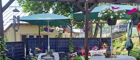 Een terras buiten met tafels en stoelen, omringd door potplanten en een pergola met bloeiende manden.