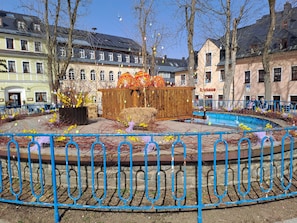 Ett offentligt torg med en fontän, en träkonstruktion med färgstarka dekorationer och byggnader i bakgrunden.