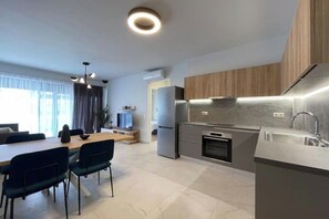 A modern kitchen with a built-in oven, a refrigerator, and a sink. There is a dining area with a table and chairs. - Athenean Oasis In Ellinikon-C1 (Elliniko)