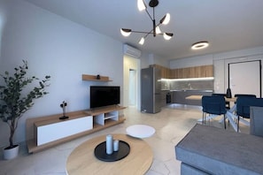 A modern living room with a flat-screen TV, a wooden entertainment unit, a dining area with blue chairs, and a kitchen in the background. - Athenean Oasis In Ellinikon-C1 (Elliniko)