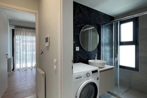 A modern bathroom with a washing machine, a sink, and a glass shower enclosure. - Athenean Oasis In Ellinikon-C1 (Elliniko)
