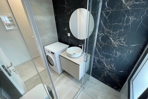 A modern bathroom with a marble wall, a round mirror, a sink, and a washing machine. - Athenean Oasis In Ellinikon-C1 (Elliniko)