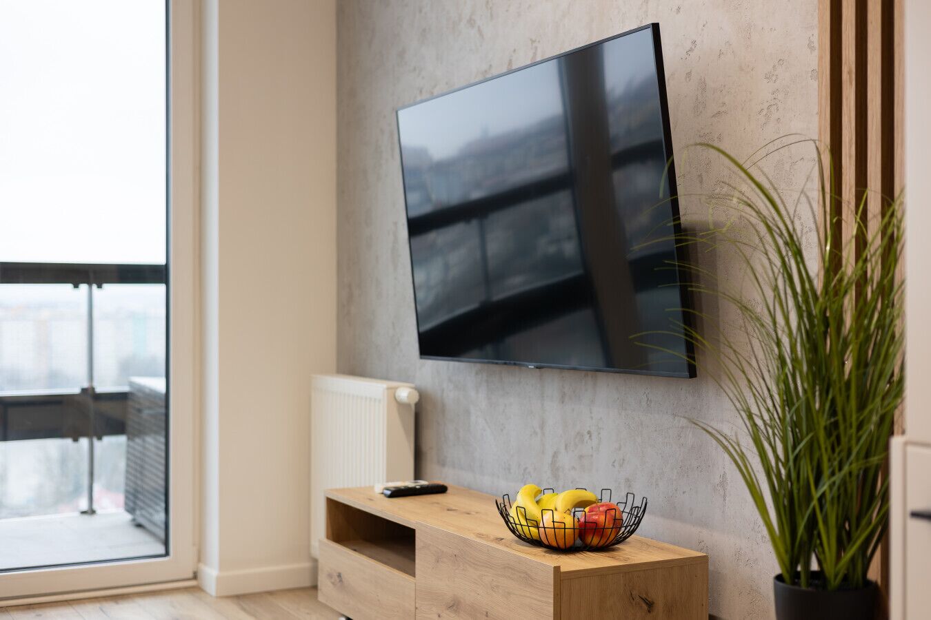 Un salon moderne avec un téléviseur à écran plat fixé sur un mur texturé, une table d’appoint en bois sur laquelle se trouve un panier de fruits, un radiateur et une grande fenêtre.