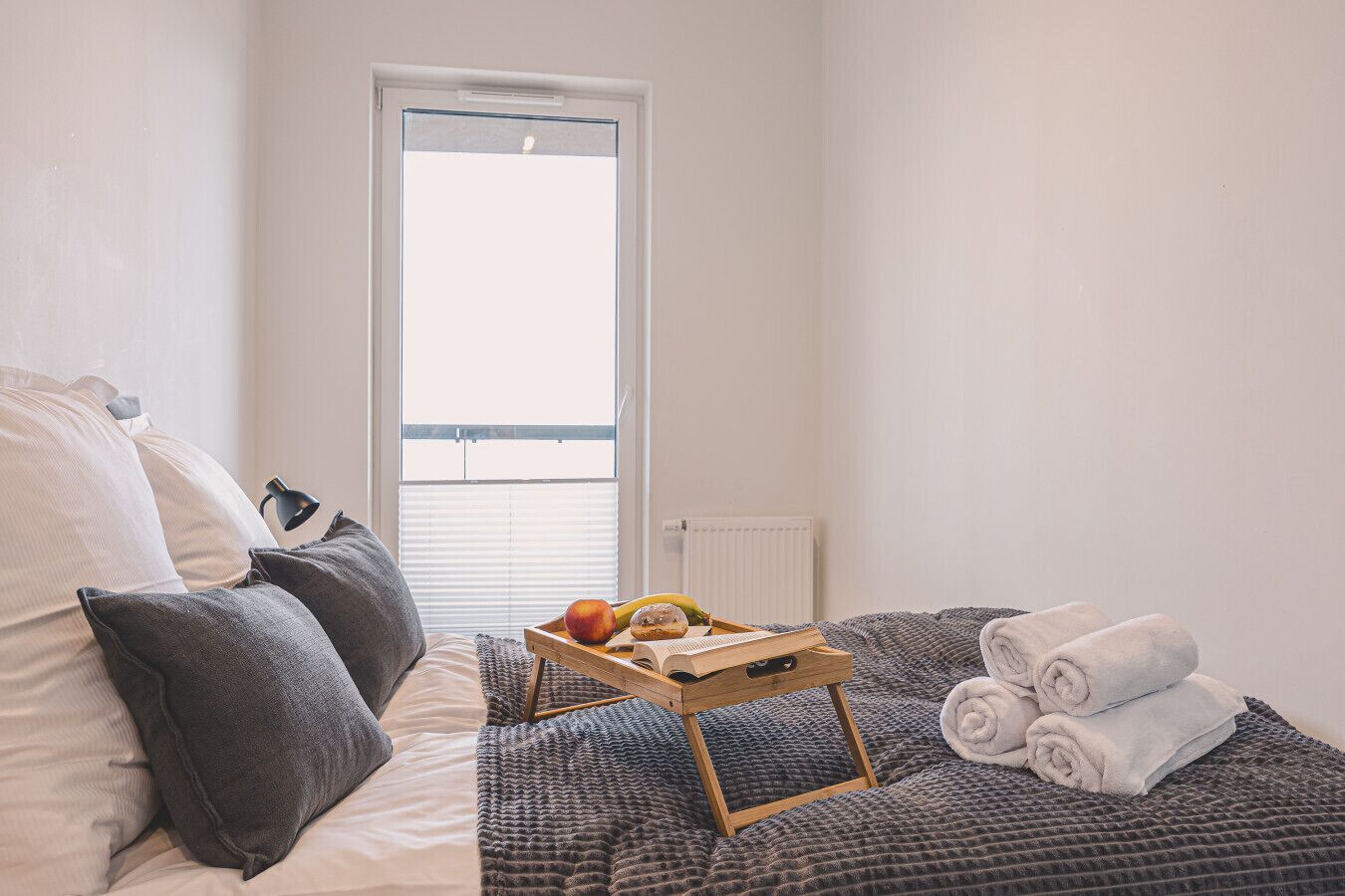 Een slaapkamer met een bed, kussens, een dienblad met fruit en een boek, een raam en een radiator.