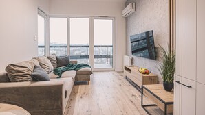 Un salon moderne avec un canapé, une table basse, un téléviseur fixé au mur et une grande fenêtre donnant sur la ville.