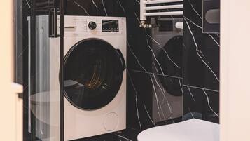 A compact laundry room with a front-loading washing machine, a toilet, and a glass partition.