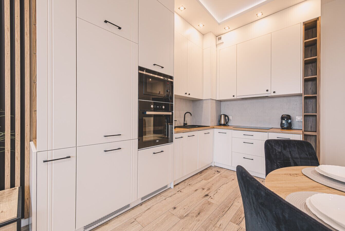 A modern kitchen with white cabinets, a built-in oven, and a dining area with a wooden table and chairs.