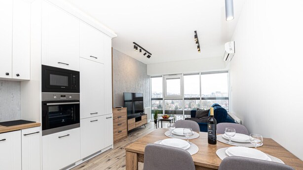 Une cuisine moderne dotée d’appareils électroménagers intégrés, un coin repas avec une table et des chaises en bois, et une grande fenêtre offrant une vue sur la ville.