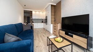 Un salon moderne avec un canapé bleu, un téléviseur posé sur un meuble en bois, et une cuisine en arrière-plan.