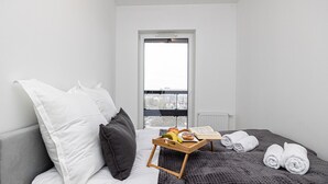 Une chambre d’hôtel moderne avec un lit, des oreillers, un plateau avec des fruits et un livre, et une vue à travers une grande fenêtre.