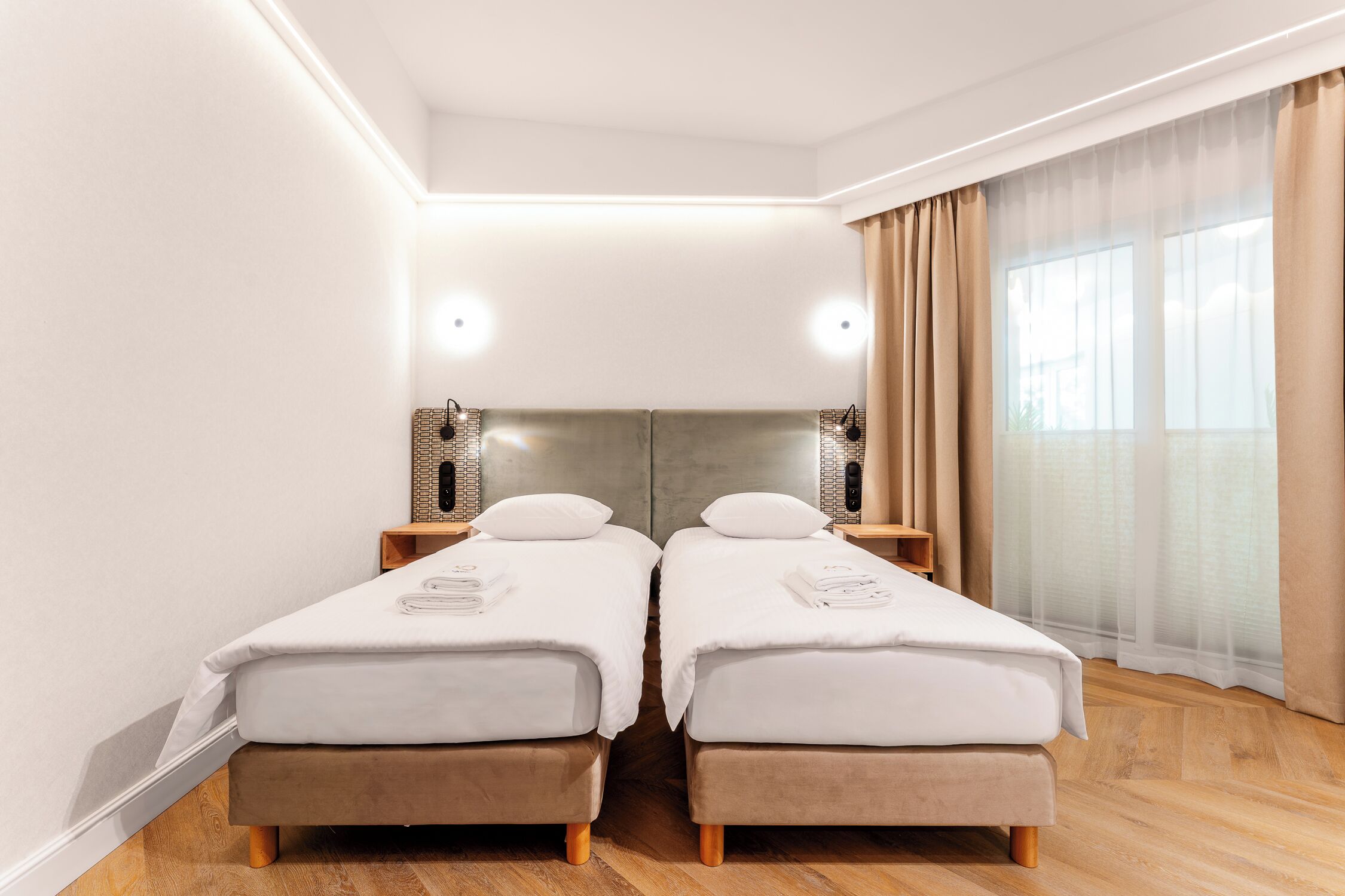 Een tweeepersoonskamer met twee bedden, een groot raam met dunne gordijnen, een houten vloer en een moderne lamp.