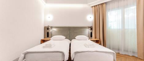 A twin room with two beds, a large window with sheer curtains, a wooden floor, and a modern light fixture.