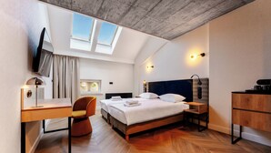 A modern hotel room with a loft-style ceiling, a bed, a desk, a chair, a wardrobe, and a small bedside table.
