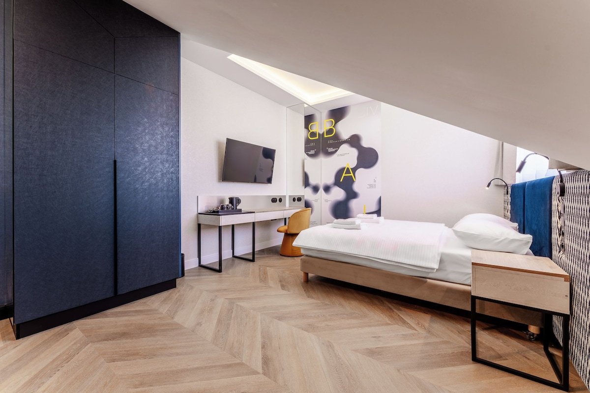 A modern hotel room with a bed, desk, chair, and a large wall art piece.